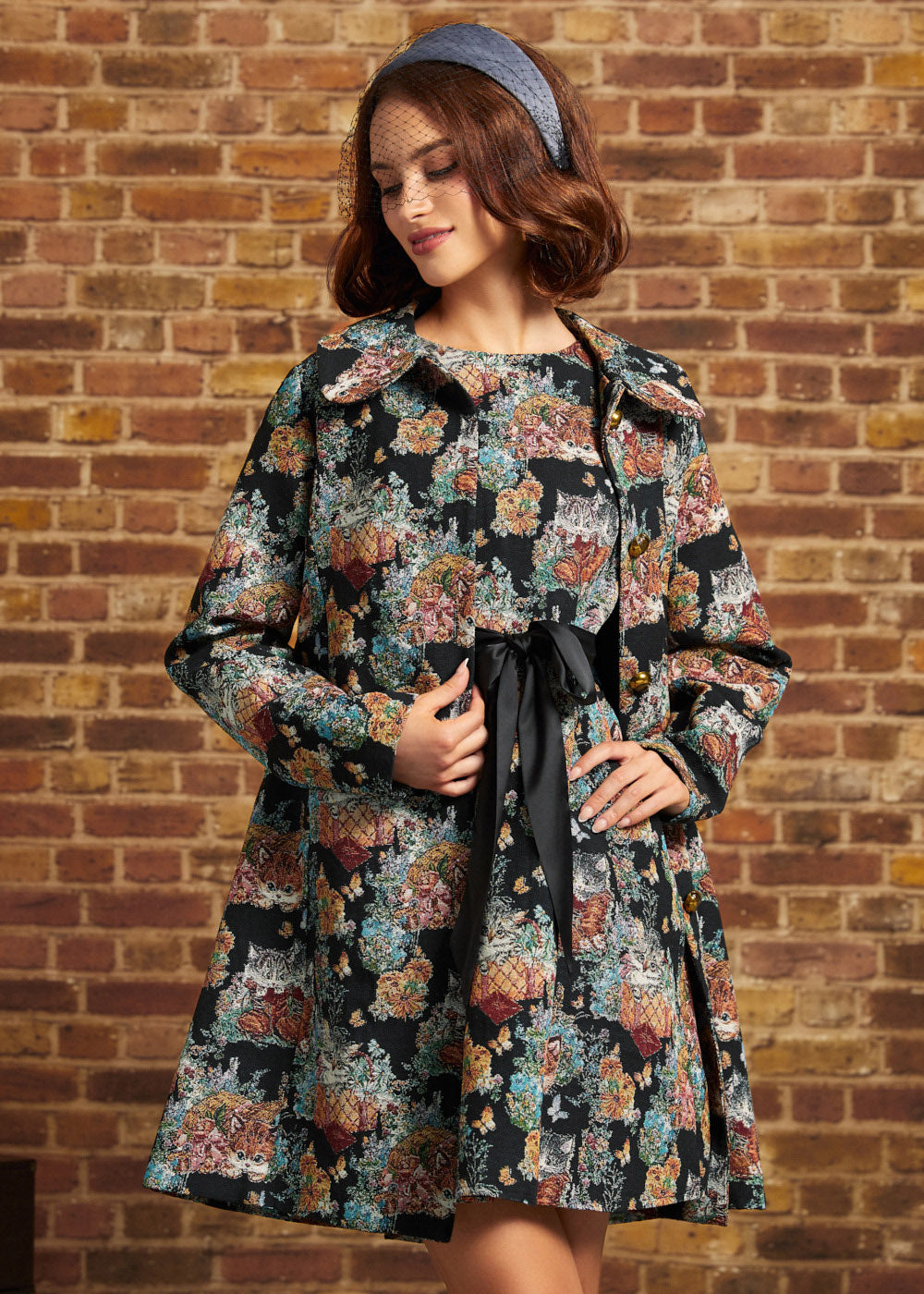 Model wearing ModCloth Timeless Brocade Midi Coat with colorful floral and cat tapestry design, gold buttons and oversized collar, styled with a blue headband