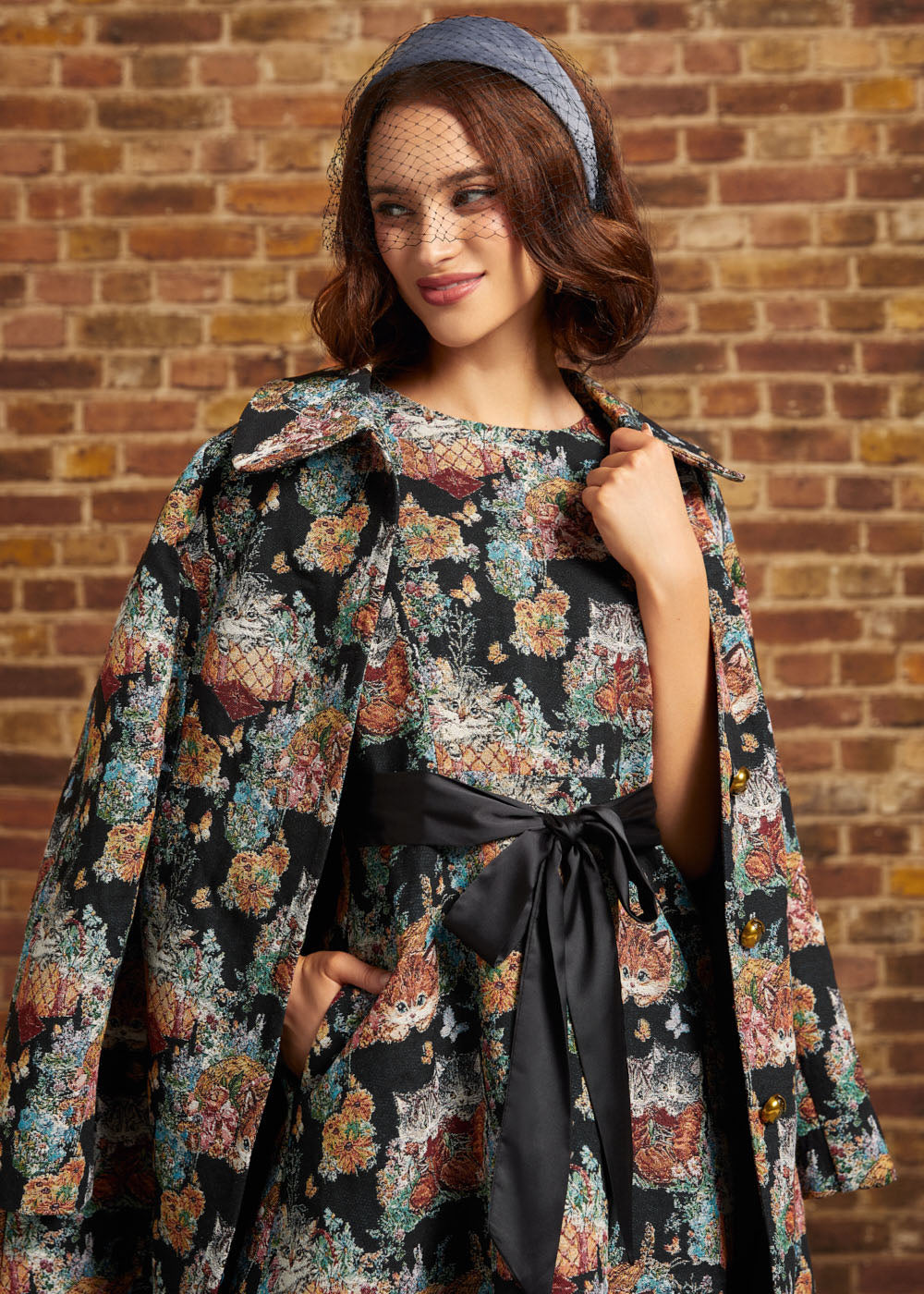 A woman wearing a black floral coat draped over her shoulders, with a matching dress with a black ribbon sash and a blue headnet, posing against a brick wall.