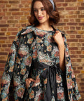 A woman wearing a black floral coat draped over her shoulders, with a matching dress with a black ribbon sash and a blue headnet, posing against a brick wall.