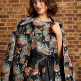 A woman wearing a black floral coat draped over her shoulders, with a matching dress with a black ribbon sash and a blue headnet, posing against a brick wall.