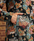 Close-up of model's hand inside side pocket of black floral cat tapestry coat, showing woven-style fabric texture and gold buttons.