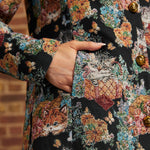Close-up of model's hand inside side pocket of black floral cat tapestry coat, showing woven-style fabric texture and gold buttons.