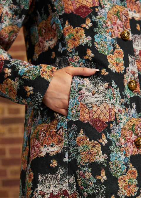 Close-up of model's hand inside side pocket of black floral cat tapestry coat, showing woven-style fabric texture and gold buttons.