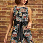 A woman wearing a sleeveless black fit-and-flare dress with a colorful floral cat print and a black satin waist tie, standing against a brick wall.