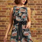A woman wearing a sleeveless black fit-and-flare dress with a colorful floral cat print and a black satin waist tie, standing against a brick wall.