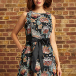 A woman models a sleeveless black fit-and-flare dress with a colorful floral print and a large black ribbon tie at the waist, standing against a brick wall background.