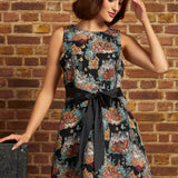 A woman models a sleeveless black A-line dress with a colorful floral cats print and a wide black ribbon tie at the waist, posed against a brick wall.