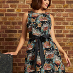 Model wearing the Tallulah sleeveless mini dress with colorful tapestry-style kitten and floral motifs, black bow-tie waist, and flared skirt against a brick wall.
