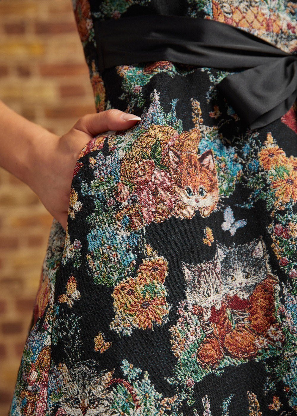 Close-up of the black tapestry-style dress fabric featuring orange and gray kittens, colorful flowers, and a model's hand resting inside a side pocket.