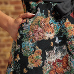 Close-up of the black tapestry-style dress fabric featuring orange and gray kittens, colorful flowers, and a model's hand resting inside a side pocket.