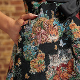 Close-up of the black tapestry-style dress fabric featuring orange and gray kittens, colorful flowers, and a model's hand resting inside a side pocket.