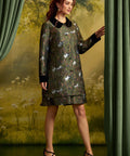 A woman models a knee-length patterned dress with a dark green base, white cats, and small floral details, featuring a black collar and black cuffs. She is posed between green curtains against a soft, painterly forest backdrop.