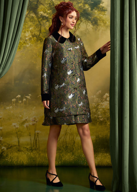 A woman models a knee-length patterned dress with a dark green base, white cats, and small floral details, featuring a black collar and black cuffs. She is posed between green curtains against a soft, painterly forest backdrop.