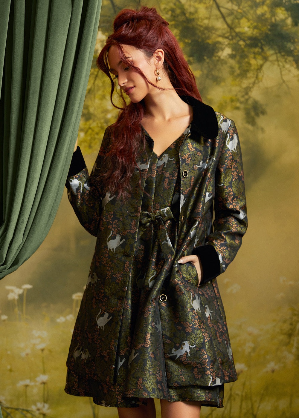 A woman with red hair wearing a dark green patterned coat with black collar and cuffs, standing in front of a green curtain and a blurred outdoor background.