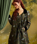 A woman with red hair wearing a dark green patterned coat with black collar and cuffs, standing in front of a green curtain and a blurred outdoor background.