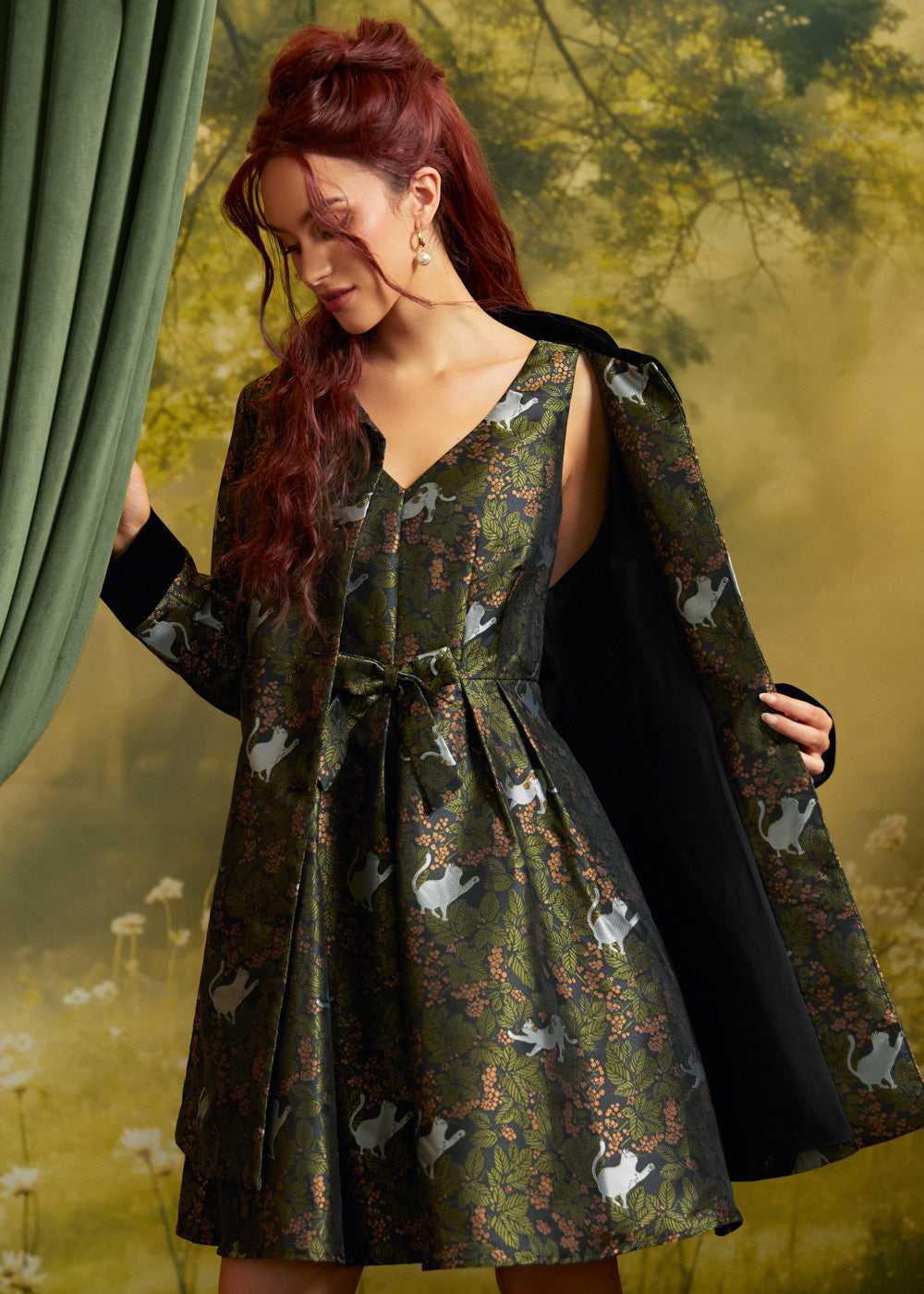 Model wearing an olive-green cat print fit-and-flare dress with its matching printed coat draped over her shoulders, showing the coordinated woodland cat pattern.