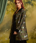 A woman models a dark green jacquard jacket featuring a white cat and floral pattern, styled over a black dress against a soft-focus forest backdrop.