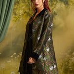 A woman models a dark green jacquard jacket featuring a white cat and floral pattern, styled over a black dress against a soft-focus forest backdrop.