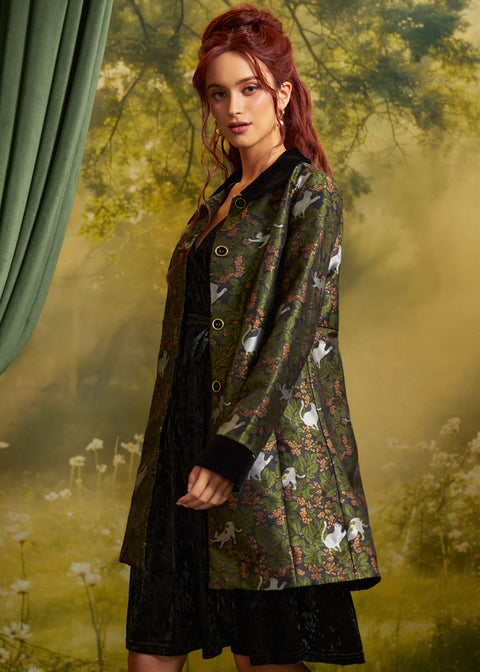 A woman models a dark green jacquard jacket featuring a white cat and floral pattern, styled over a black dress against a soft-focus forest backdrop.