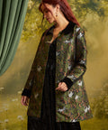 A woman with red hair models a green patterned coat with black collar and cuffs, featuring a cat print, over a black dress against a soft, misty forest backdrop.