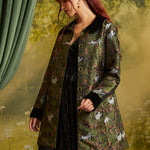A woman with red hair models a green patterned coat with black collar and cuffs, featuring a cat print, over a black dress against a soft, misty forest backdrop.