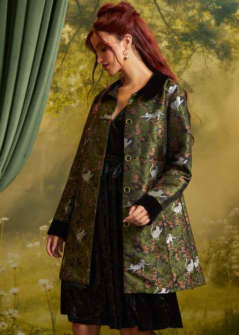 A woman with red hair models a green patterned coat with black collar and cuffs, featuring a cat print, over a black dress against a soft, misty forest backdrop.