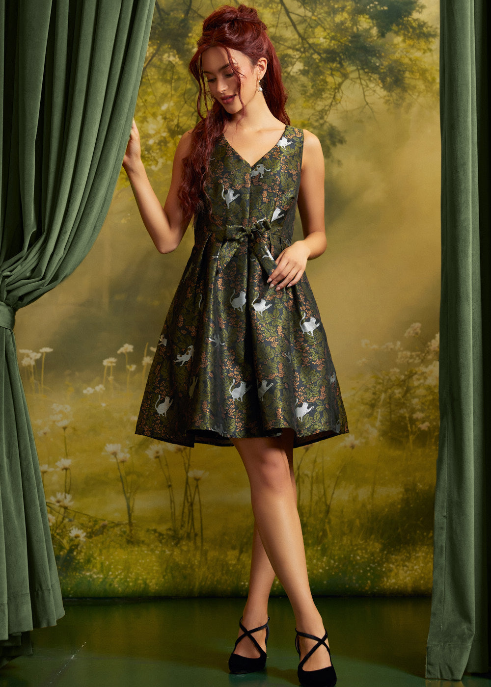 A woman models a sleeveless V-neck A-line dress with a dark green and black floral pattern featuring white cats, posed between green curtains against a forest backdrop.