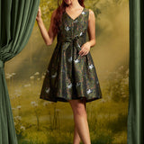 A woman models a sleeveless V-neck A-line dress with a dark green and black floral pattern featuring white cats, posed between green curtains against a forest backdrop.