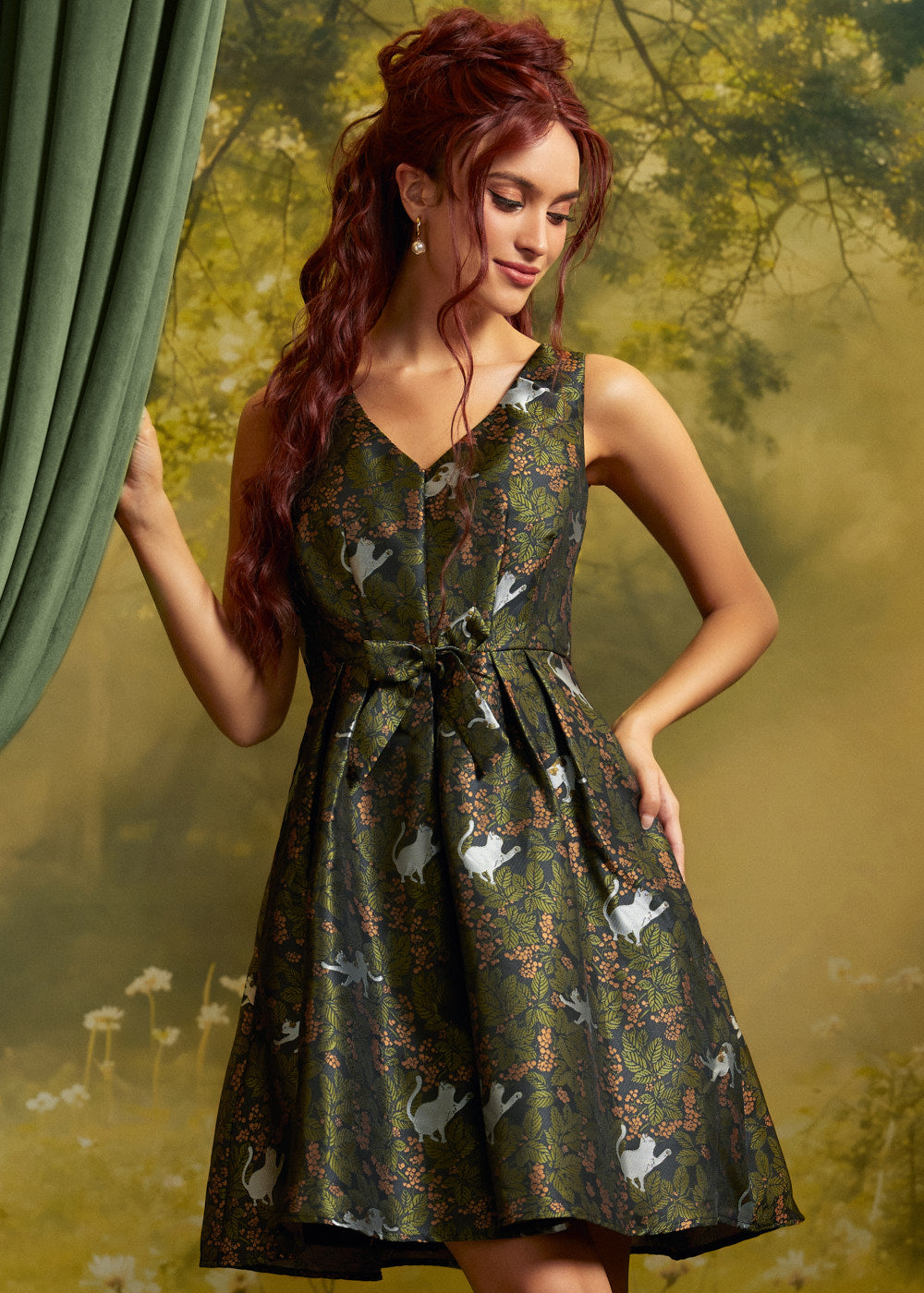 A woman with long red hair models a sleeveless V-neck mini dress with a green and black animal print featuring white cats. She is posed in front of a soft-focus forest background with a green curtain on the left.