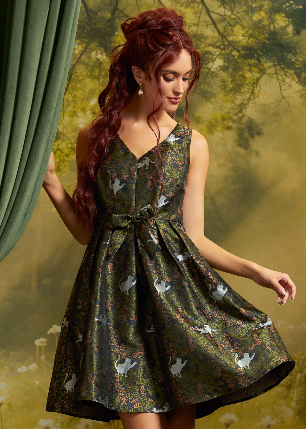 Model wearing a sleeveless olive-green cat print fit-and-flare dress with V-neckline, matching waist bow and pleated skirt, holding the skirt out against a woodland backdrop.