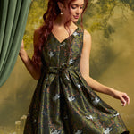 Model wearing a sleeveless olive-green cat print fit-and-flare dress with V-neckline, matching waist bow and pleated skirt, holding the skirt out against a woodland backdrop.