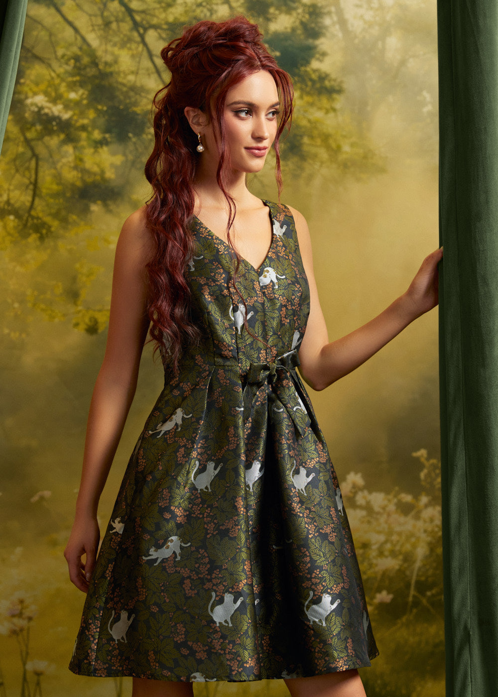 A woman with long red hair models a sleeveless V-neck mini dress with a dark green satin-like finish and an allover print of white cats and pink floral details. She is posed against a soft-focus forest backdrop with a green curtain on the right.