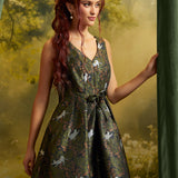 A woman with long red hair models a sleeveless V-neck mini dress with a dark green satin-like finish and an allover print of white cats and pink floral details. She is posed against a soft-focus forest backdrop with a green curtain on the right.