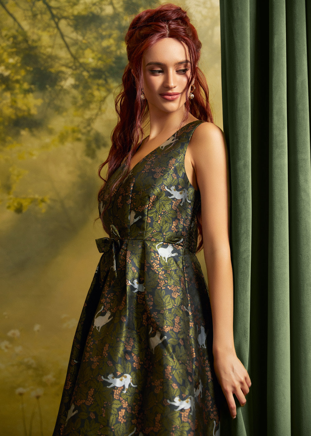 A woman wearing a sleeveless V-neck midi dress with a dark green crisscross jacquard pattern featuring white cats and orange floral details, posed next to a dark green curtain.