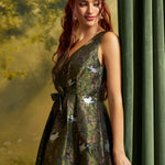 A woman wearing a sleeveless V-neck midi dress with a dark green crisscross jacquard pattern featuring white cats and orange floral details, posed next to a dark green curtain.