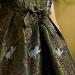 Close-up of the olive-green botanical dress showing white cat illustrations, leafy foliage, copper-colored blossoms, and front bow.