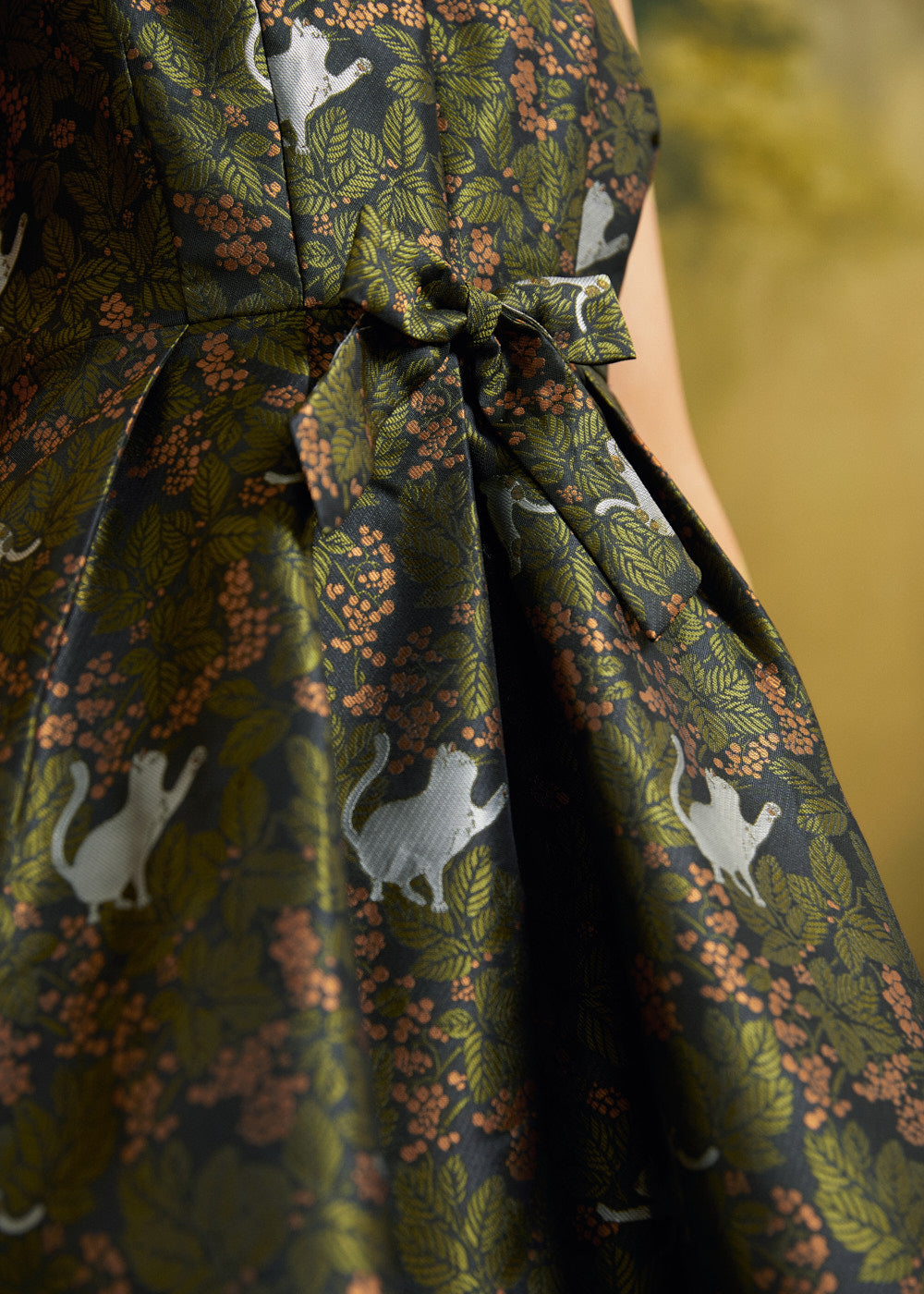 Close-up of the olive-green botanical dress showing white cat illustrations, leafy foliage, copper-colored blossoms, and front bow.