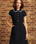 A woman wearing a black short-sleeve dress with a white lace trimmed Peter Pan collar and white button details, standing against a rustic brick wall.