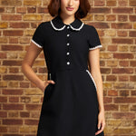 A woman wearing a black short-sleeve dress with a white lace trimmed Peter Pan collar and white button details, standing against a rustic brick wall.