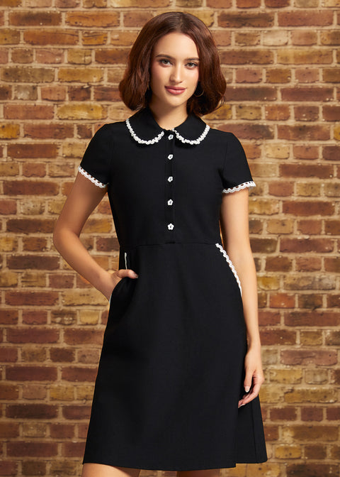 A woman wearing a black short-sleeve dress with a white lace trimmed Peter Pan collar and white button details, standing against a rustic brick wall.