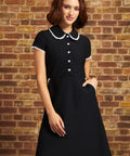 A woman wearing a black short-sleeve dress with a Peter Pan collar and white button details, standing against a brown brick wall.