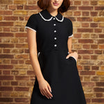 A woman wearing a black short-sleeve dress with a Peter Pan collar and white button details, standing against a brown brick wall.