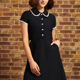 A woman wearing a black short-sleeve dress with a Peter Pan collar and white button details, standing against a brown brick wall.
