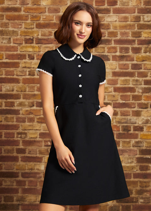 A woman wearing a black short-sleeve dress with a Peter Pan collar and white button details, standing against a brown brick wall.