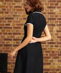 A woman wearing a short-sleeve black dress with white lace trim on the collar and sleeve cuffs, photographed from the back against a rustic brick wall.