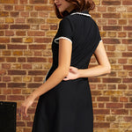 A woman wearing a short-sleeve black dress with white lace trim on the collar and sleeve cuffs, photographed from the back against a rustic brick wall.