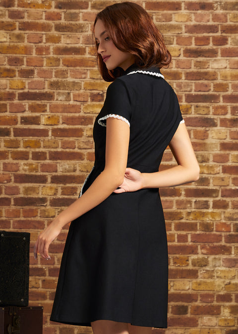 A woman wearing a short-sleeve black dress with white lace trim on the collar and sleeve cuffs, photographed from the back against a rustic brick wall.