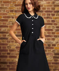 Model standing with hands in the lace-trimmed side pockets of the Lottie black fit-and-flare dress, highlighting its flower-button bodice and flared skirt.