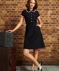 A woman models a black short-sleeve A-line dress with a white lacePeter Pan collar and white buttons down the front, standing in front of a brick wall next to vintage stacked trunks.
