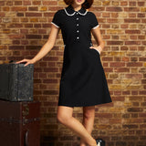 A woman models a black short-sleeve A-line dress with a white lacePeter Pan collar and white buttons down the front, standing in front of a brick wall next to vintage stacked trunks.